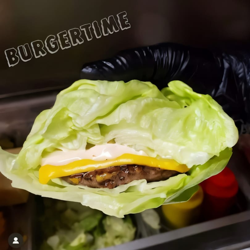A gloved hand holds a Burger Time lettuce-wrapped burger with cheese and sauce.