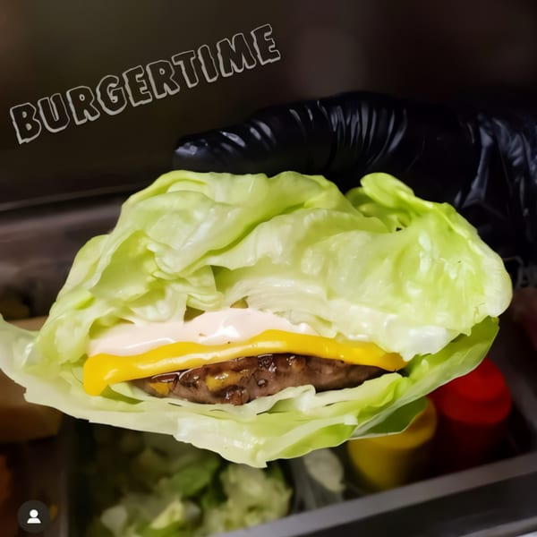 A hand in a black glove holds a Burger Time lettuce-wrapped burger with cheese and sauce.