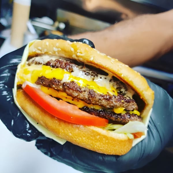 Hands in black gloves holding a Burger Time double cheeseburger with tomato, lettuce, and melted cheese.