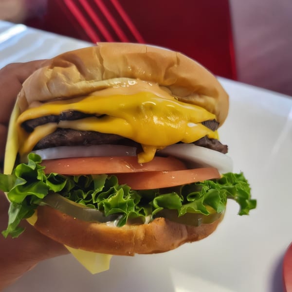 A hand holds a Burger Time double cheeseburger with melted cheese, lettuce, tomato, onion, and pickles.